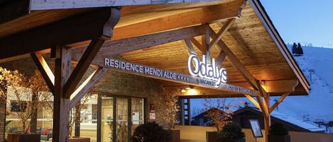 A hotel entrance with a wooden facade, signage for 'Residence Mendi Alde' and 'Hotel Le Chamois', and a view of the snowy mountain in the background.