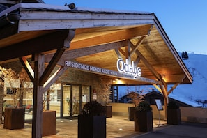 A hotel entrance with a wooden facade, signage for 'Residence Mendi Alde' and 'Hotel Le Chamois', and a view of the snowy mountain in the background. - Hôtel et Spa Le Chamois by Odalys (La Clusaz)