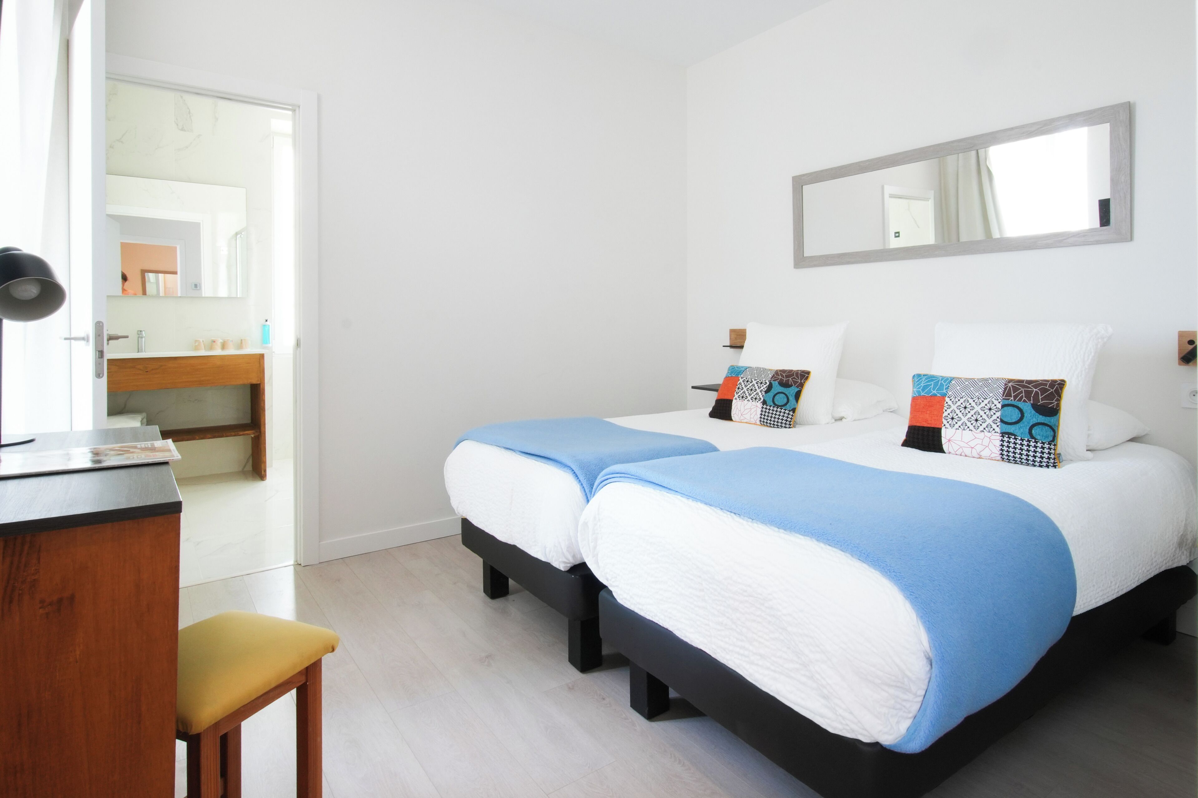 Un dormitorio con dos camas, un escritorio de madera, una silla y un baño visible a través de una puerta abierta.