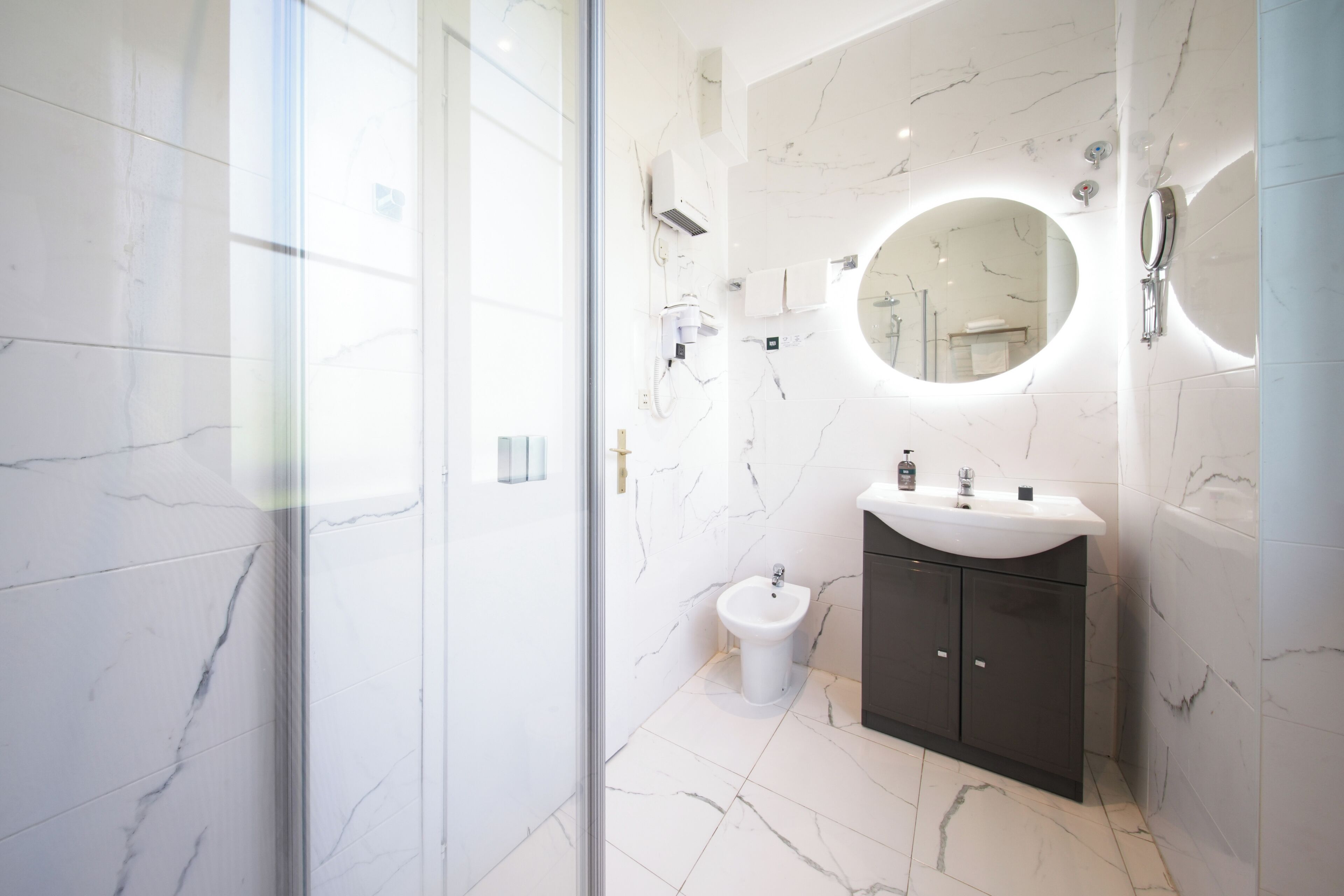 Un baño moderno con piso de mármol, un espejo redondo, un lavamanos y un bidé.