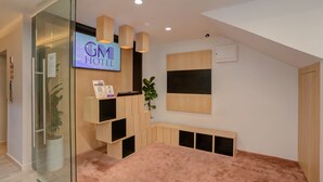A hotel room with a glass door entrance, a reception desk, a television displaying the hotel name, a plant, a wooden bench, and a large window. - GM Hotel Sunway Metro (Petaling Jaya)