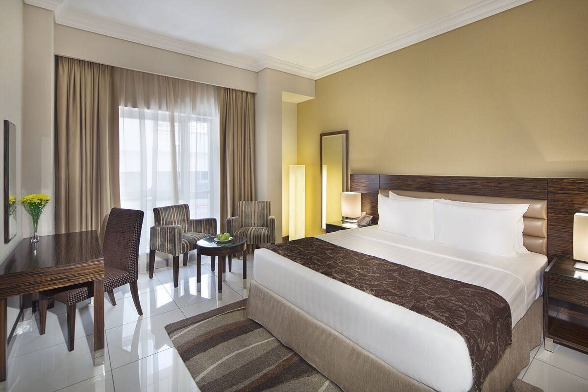 A hotel room with a large bed, a desk with a chair, a small table with a vase of flowers, and a window with curtains.