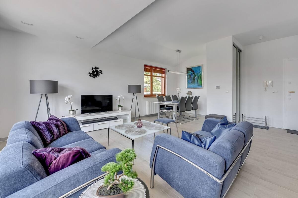 Ein modernes Wohnzimmer mit Flachbildfernseher, einem Essbereich mit Tisch und Stühlen und einem großen Fenster mit Jalousien.