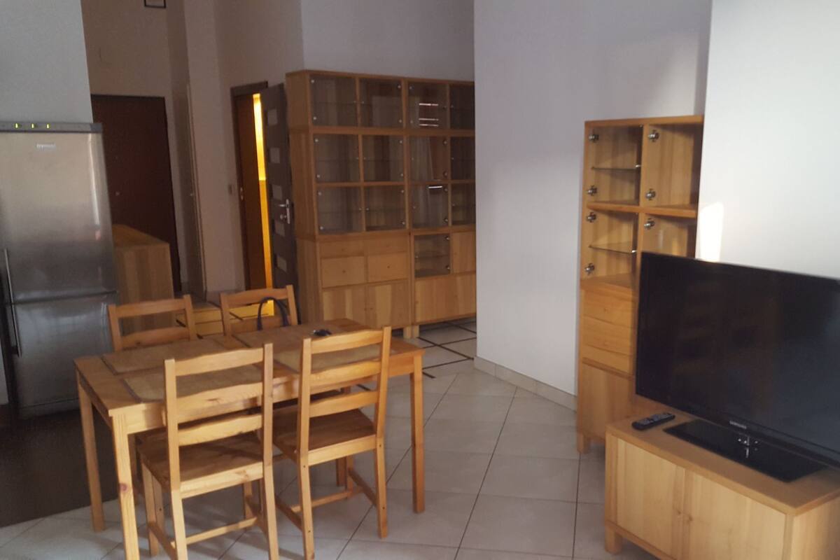 Ein gemütlicher Wohnraum mit Esstisch aus Holz, Stühlen, einem Fernseher auf einem Holzständer und einer eingebauten Holzvitrine mit Glastüren.