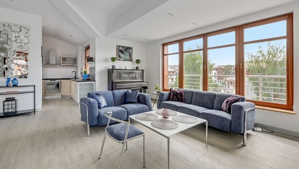 Un salón moderno con un sofá gris, una mesa de centro y una mesa de comedor. Se observa una cocina al fondo, con un piano y un ventanal grande.