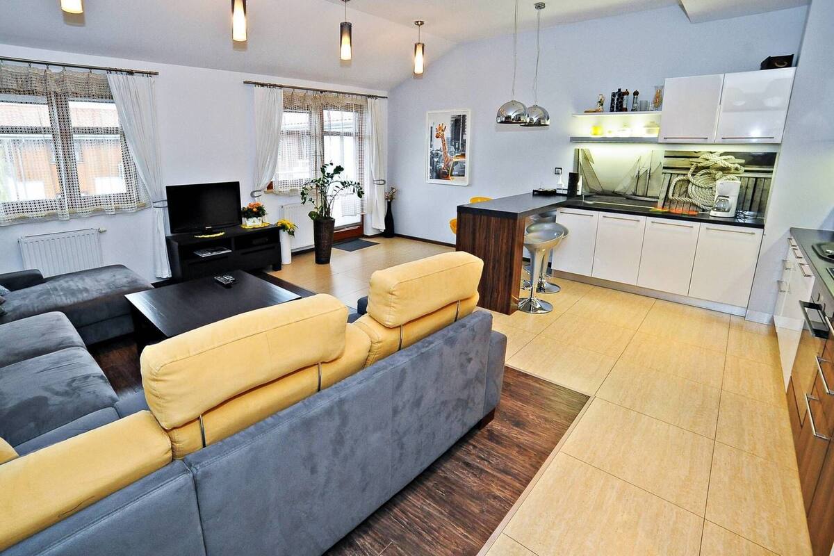 Ein modernes Wohnzimmer mit einem grauen Sofa, einer schwarzen Unterhaltungsecke und einem Essbereich, im Hintergrund ist eine Küche zu sehen.