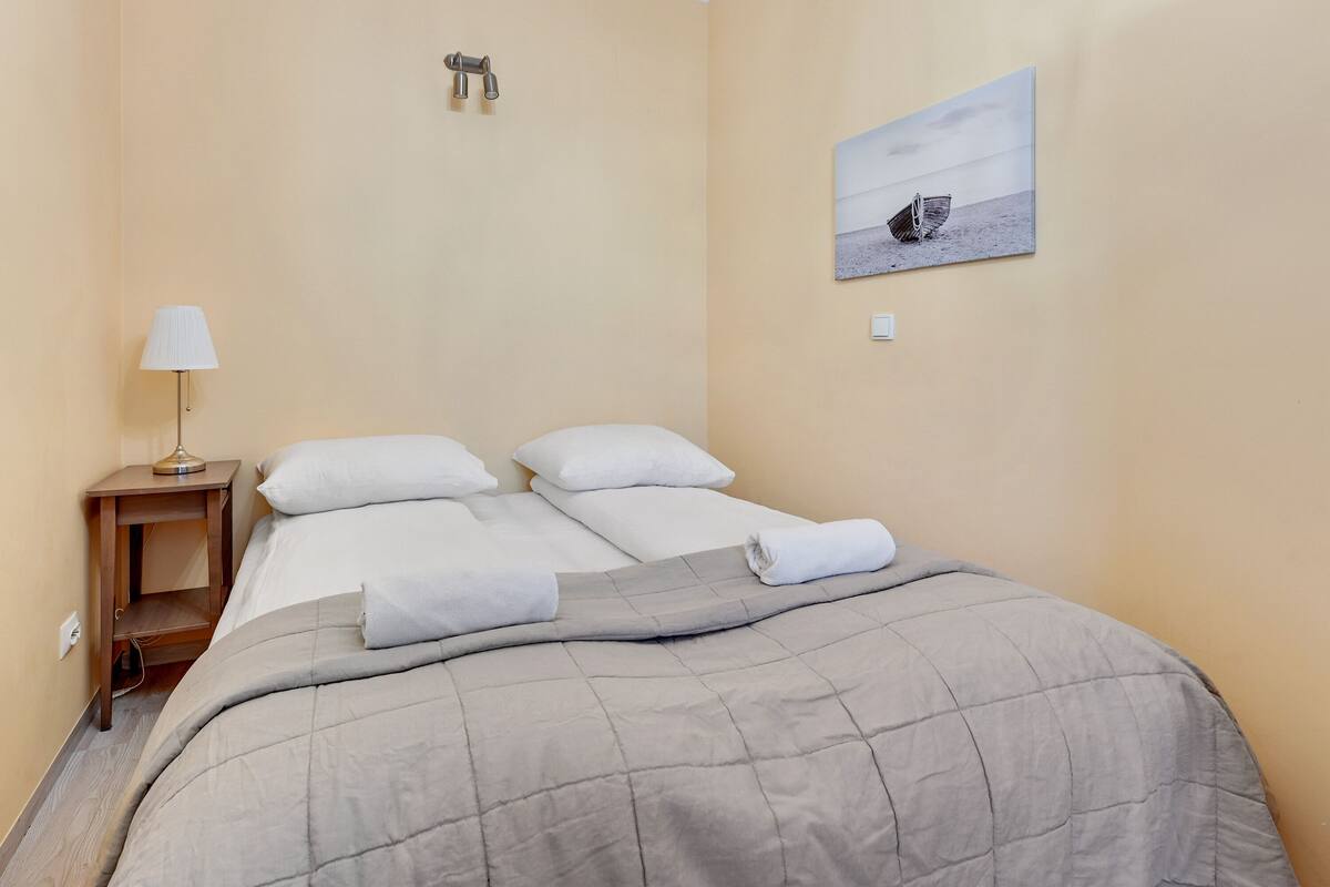 Ein ordentlich bezogenes Bett mit einer grauen Steudecke, ein Nachttisch mit einer Lampe und ein Gemälde eines Bootes an der Wand.