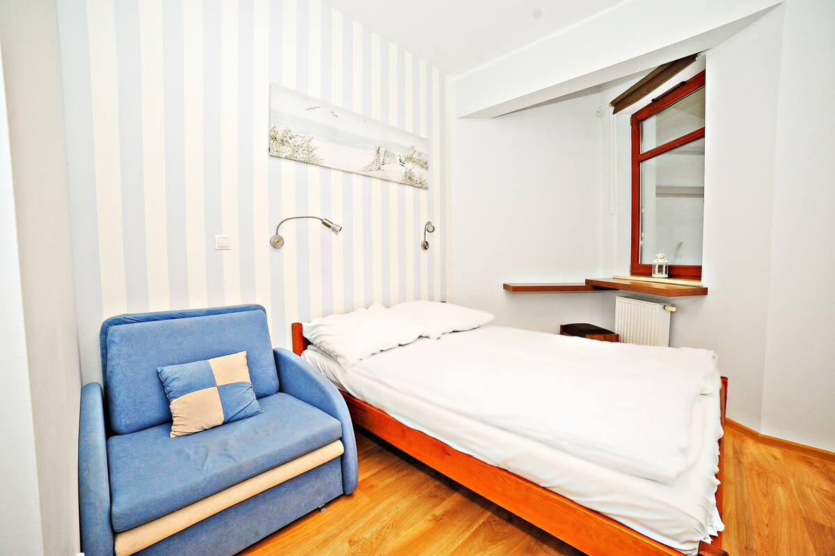 Ein Schlafzimmer mit einem Bett, einem blauen Sofa, Holzboden, Heizkörper und einem Fenster mit Holzrahmen.