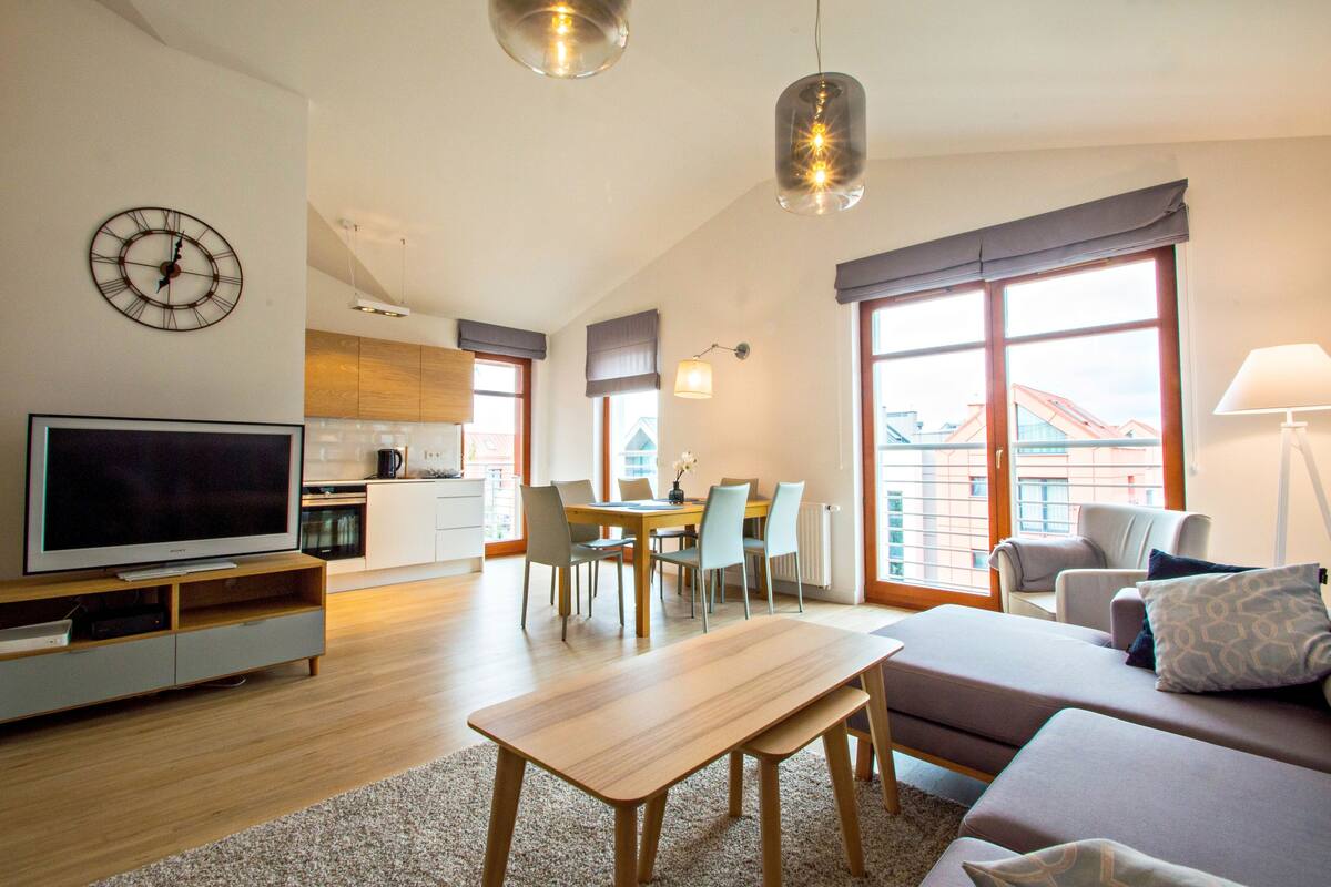 Ein modernes Wohnzimmer mit Flachbildfernseher, Couchtisch aus Holz und einer Couch mit Kissen. Es gibt eine Essecke mit Tisch und Stühlen, und im Hintergrund eine Küche.
