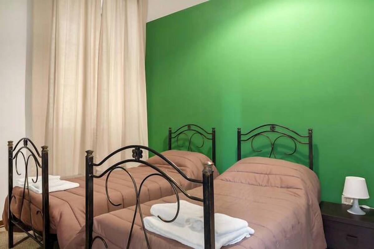 Une chambre avec deux lits, chacun doté d’un cadre en métal et de draps bruns. Un mur est peint en vert et une lampe blanche est posée sur une table de chevet.