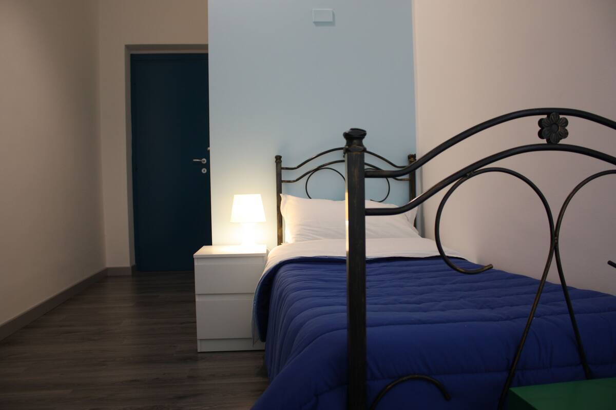 Un lit simple avec un cadre en métal et une couvre-lit bleue, une table de chevet blanche avec une lampe, et une porte fermée dans une pièce dont le sol est recouvert de parquet.