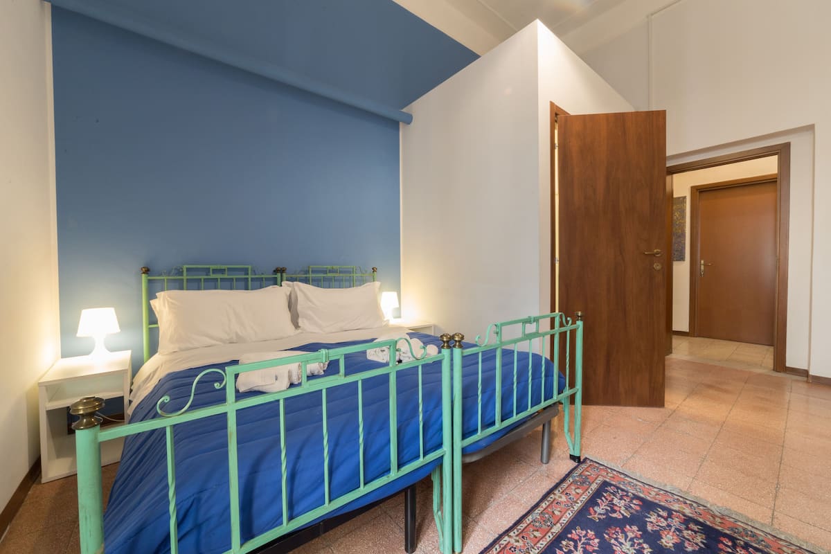 Une chambre avec un lit bleu, des oreillers blancs, un cadre de lit en métal vert, une table de chevet avec une lampe, et une porte en bois en arrière-plan.