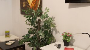 A room with a white desk, a chair, a potted plant, a painting on the wall, a television mounted above the desk, and a small table with a book and a remote control. - B&B Tra L'Etna e il Mare (Catania)