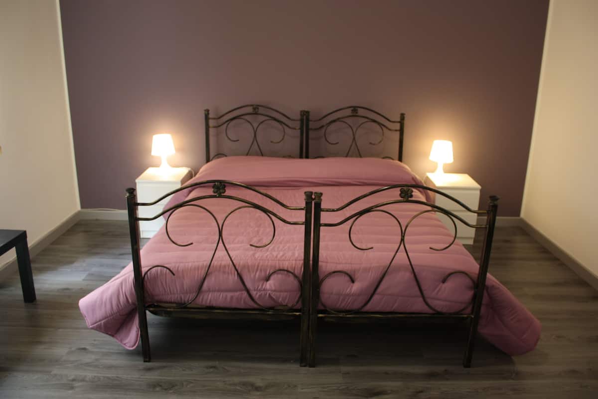 Un lit avec un cadre en métal et une couvre-lit rose, dans une pièce dont le sol est recouvert de parquet et où se trouvent deux lampes de chevet.