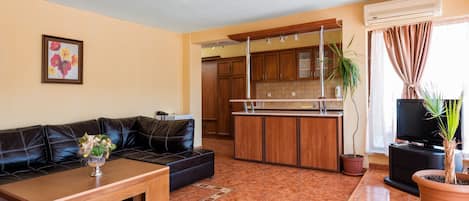 Sala de estar con sofá, mesa de centro y televisor. Se observa una zona de cocina con armarios y encimera.