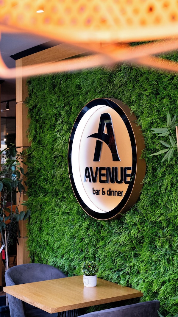 A restaurant sign with a logo and name, surrounded by a green wall and a table with chairs. - Hotel Avenue (Burgas)