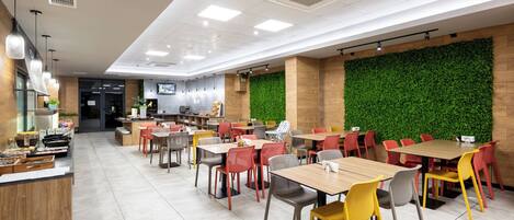 A modern restaurant with multiple dining tables and chairs, a kitchen area, and a green wall.