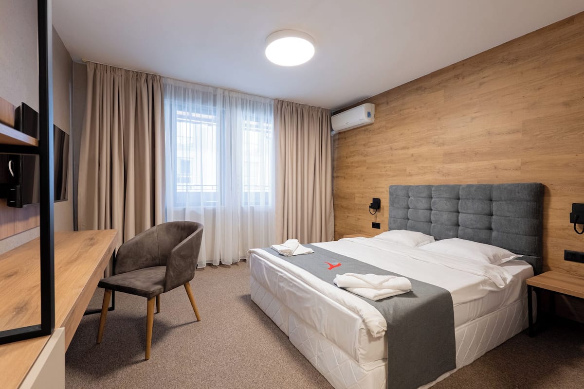 Ein modernes Hotelzimmer mit einem Holzkopfteil, einem Einzelbett, einem Schreibtisch mit Stuhl und einem großen Fenster mit Vorhängen.