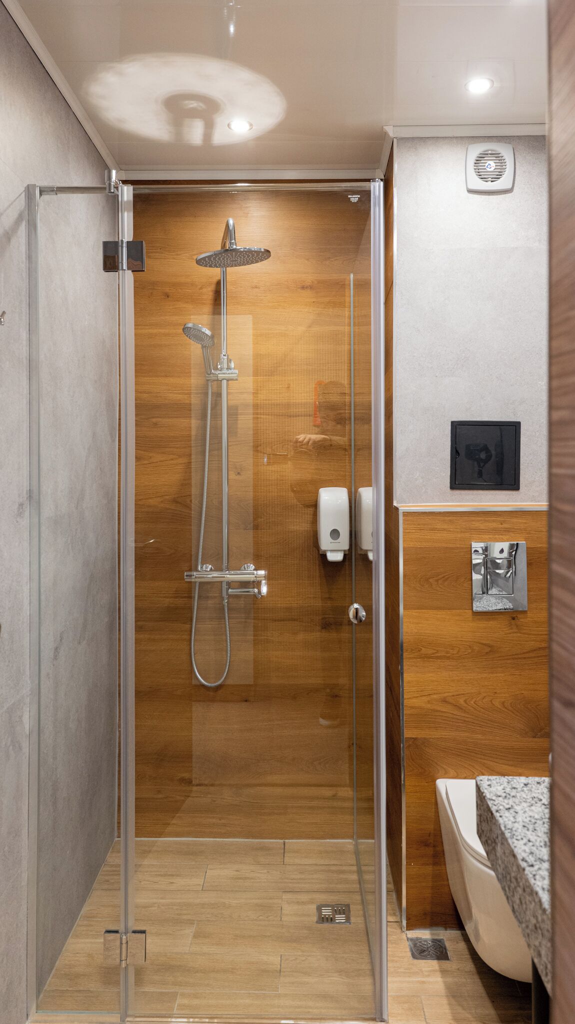 A modern shower with a glass enclosure, wooden wall paneling, and a visible hand dryer.