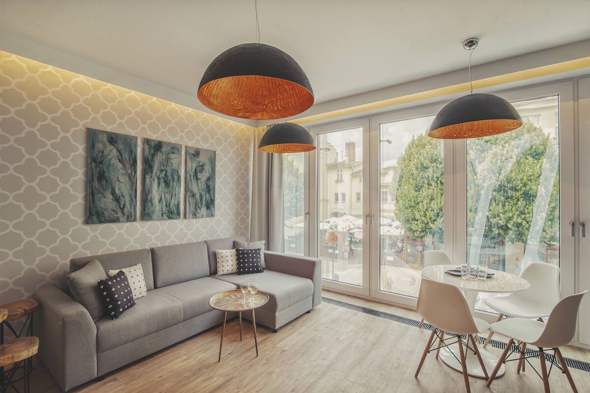 A modern living room with a grey sofa, a round coffee table, and a dining area with white chairs. There are two pendant lights hanging from the ceiling, and a large window with a view of the city.