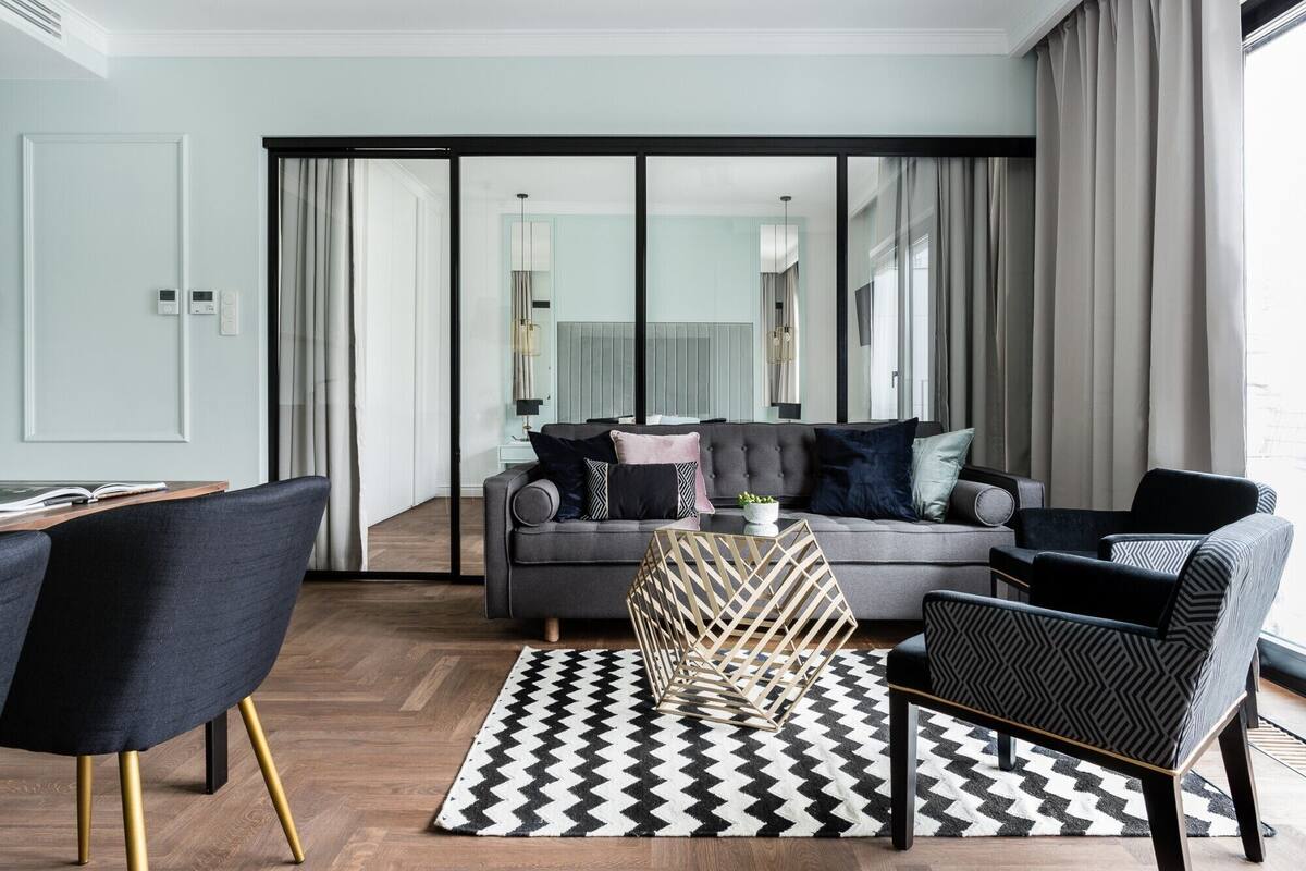 A modern living room with a grey sofa, black and white patterned rug, and a glass partition leading to a balcony.