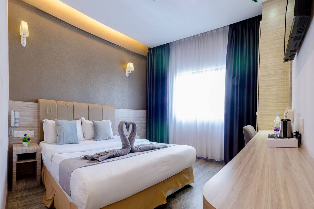 Ein Hotelzimmer mit einem großen Bett, einem Nachttisch mit Telefon, einer Lampe und einer kleinen Pflanze. Es gibt einen Schreibtisch mit Stuhl und einer Kaffeemaschine.