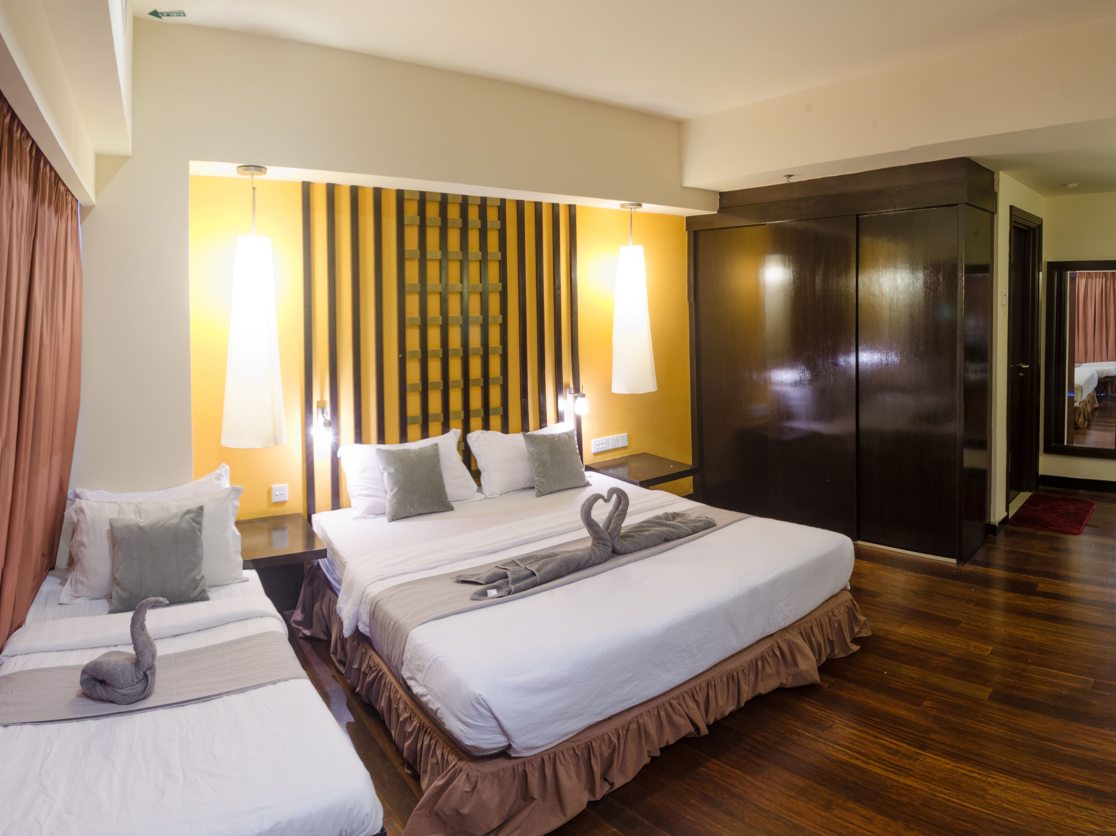 A hotel room with two beds, a wooden floor, a dark wardrobe, and a wall with vertical wooden slats and a hanging lamp.