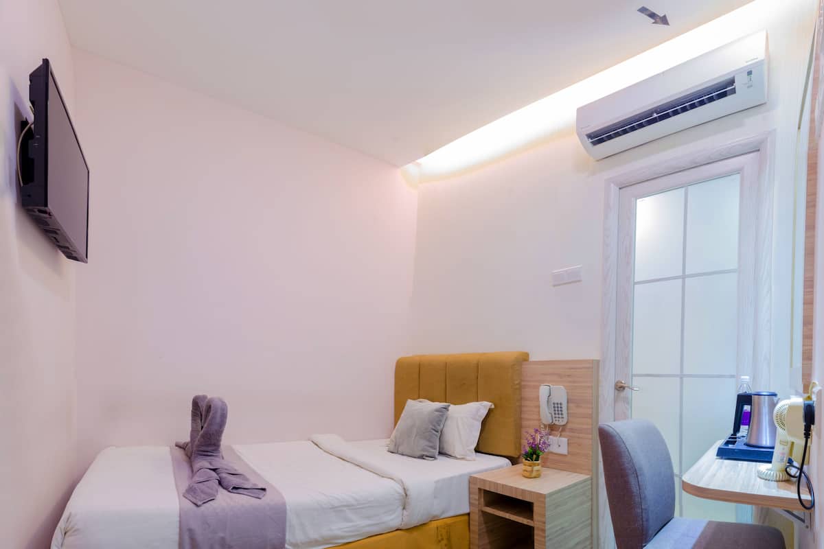 Ein kleines Hotelzimmer mit einem Einzelbett, einem Nachttisch aus Holz, einem Schreibtisch mit Stuhl und einer Klimaanlage.