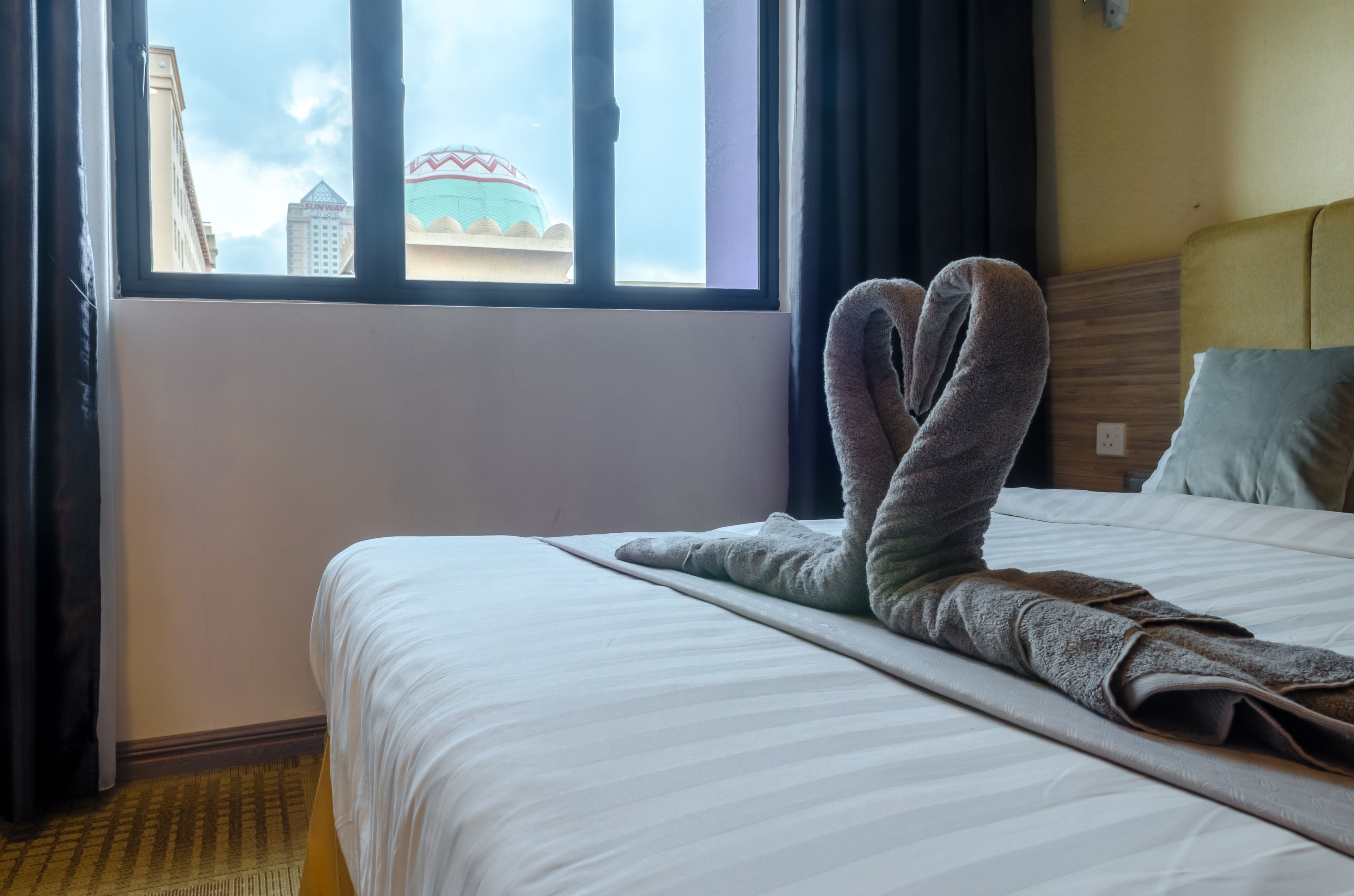 Ein Hotelzimmer mit einem Bett, einem Fenster mit Blick auf ein Gebäude und einem Handtuch auf dem Bett.