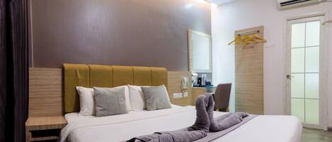 Habitación de hotel con una cama grande, cabecero de madera, mesita de noche, espejo, escritorio con silla y unidad de aire acondicionado.