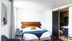 A modern bedroom with a wooden headboard, a bed with white and blue bedding, a grey armchair, and a dark wooden floor. - Hotel Napoléon (Ile-d'Aix)