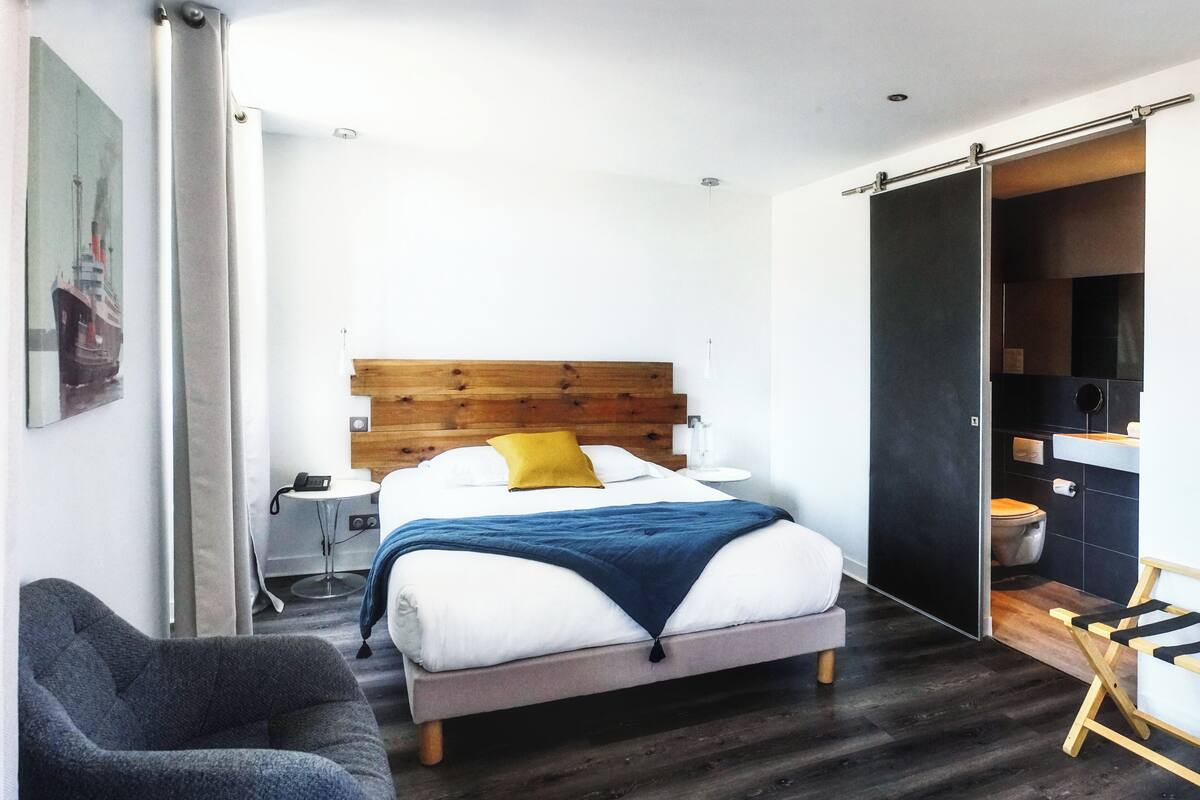 A modern bedroom with a wooden headboard, a bed with white and blue bedding, a grey armchair, and a dark wooden floor.