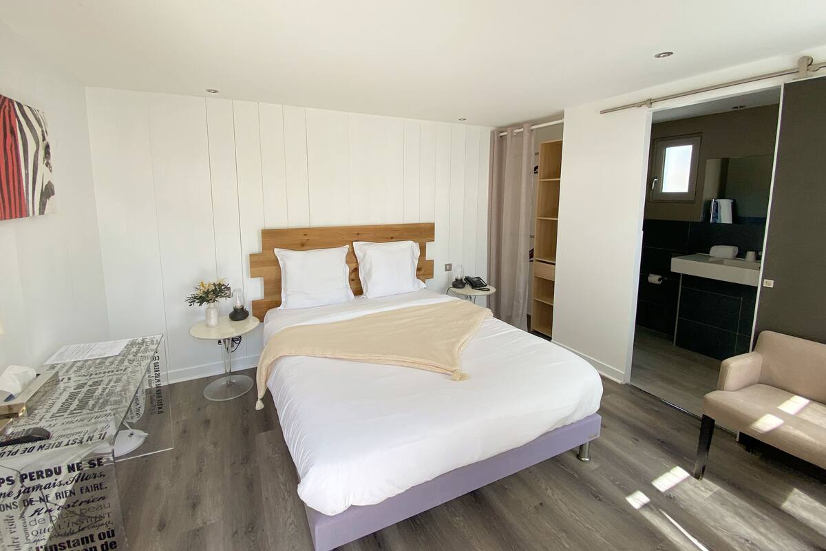 A modern hotel room with a large bed, a bedside table with a lamp, a small side table, a wardrobe, and a seating area with a chair.