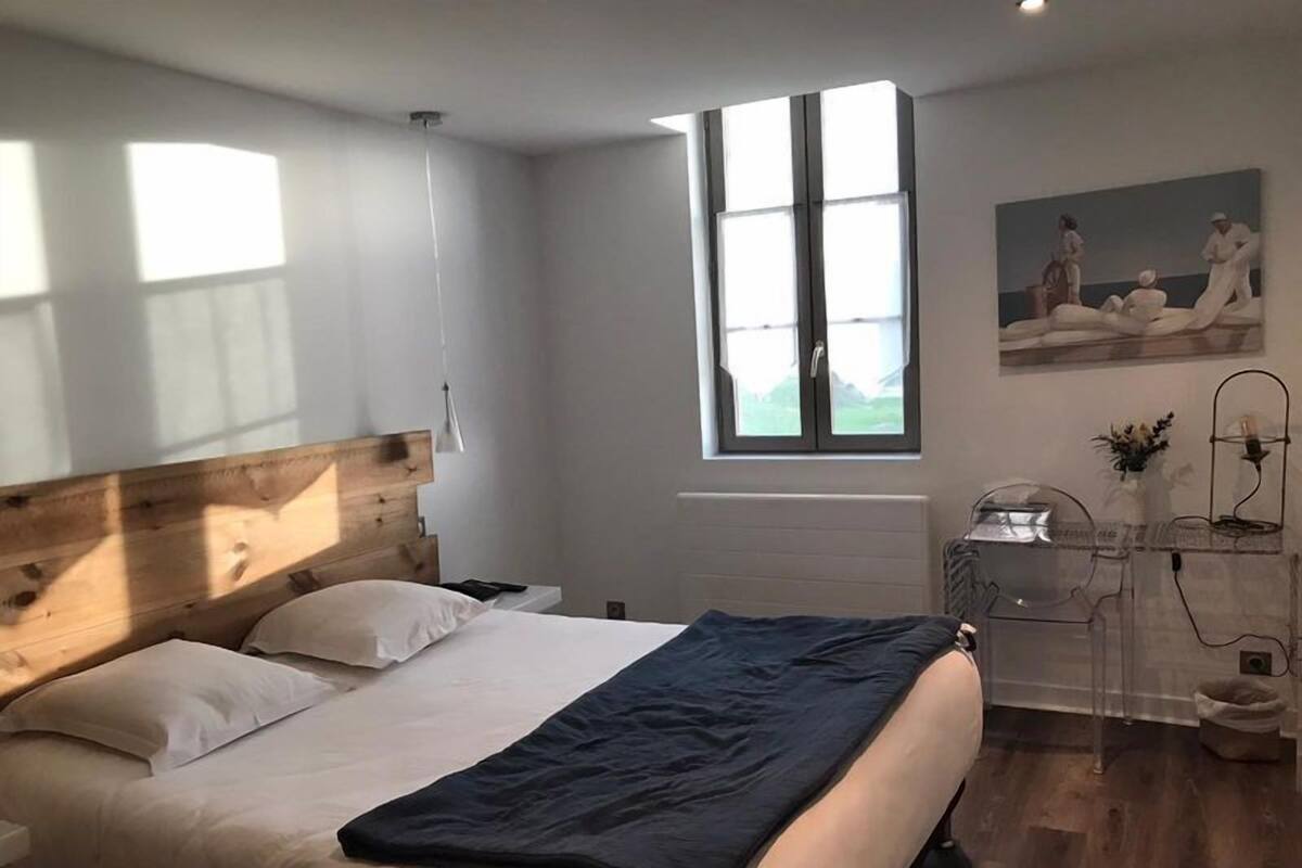 A bedroom with a wooden headboard, a large bed with white and dark blue bedding, a desk with a lamp, and a window with a view of the outdoors.