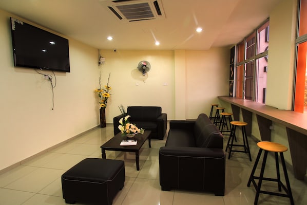 A modern waiting area with a flat-screen TV, comfortable seating, a fan, and a bar area with stools.