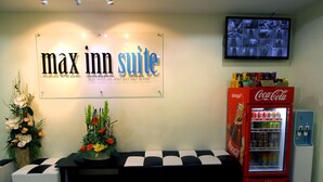 A reception area with a 'max inn suite' sign, a television, a Coca-Cola refrigerator, and a floral arrangement. - Max Inn Suite Hotel (Batu Pahat)
