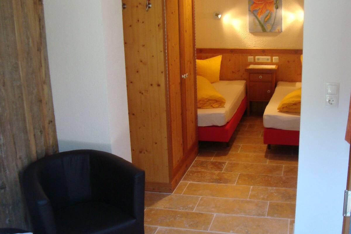 A bedroom with two beds, a wooden wardrobe, a chair, and a painting on the wall.