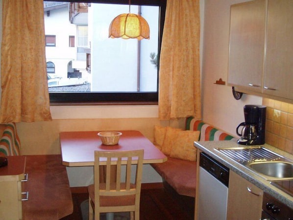 A compact kitchen with a dining table, a single chair, a coffee maker, and a window with curtains.