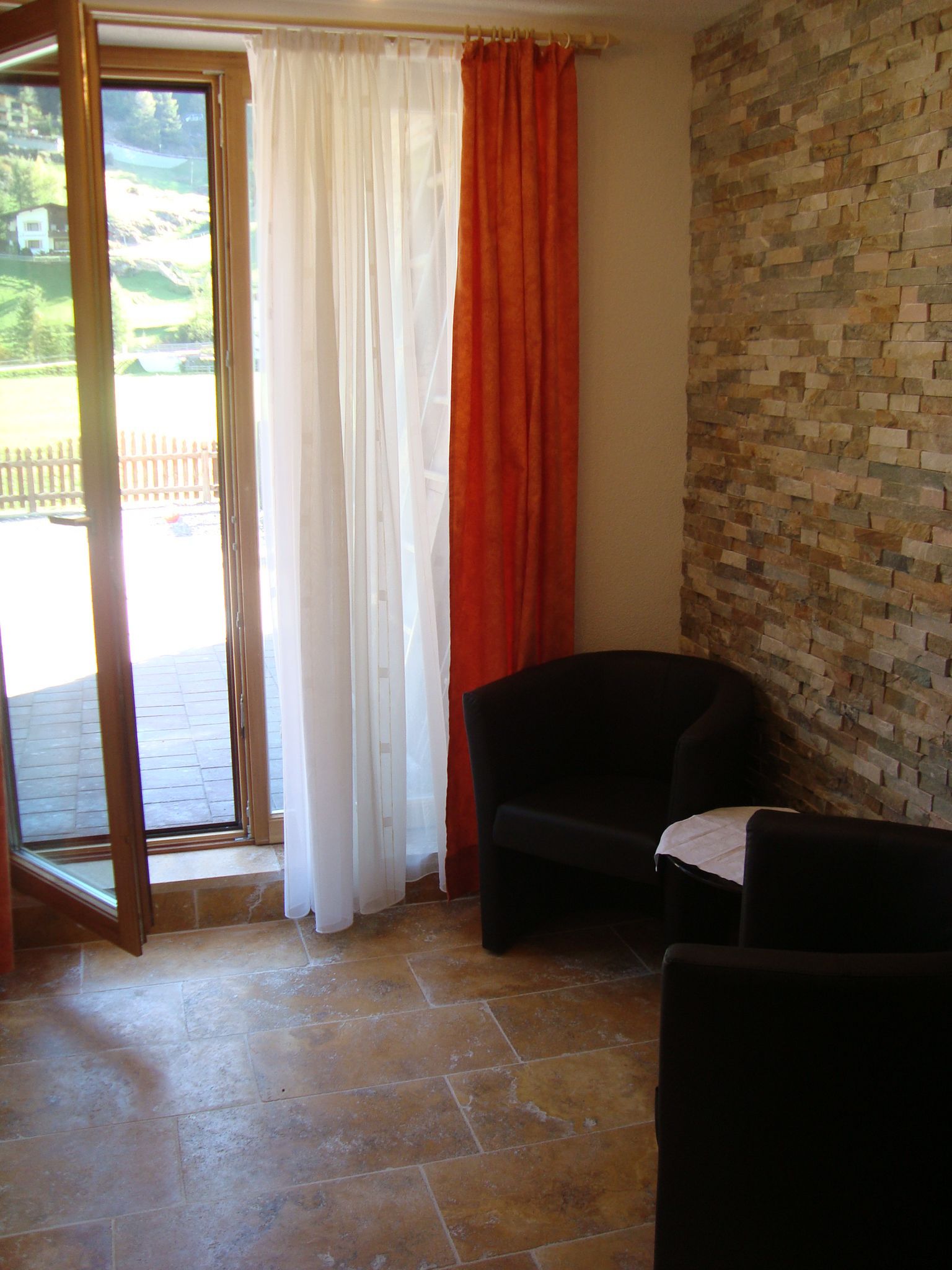 Una habitación con suelo de baldosas de piedra, una silla negra, una cortina roja y vistas a una valla de madera a través de una puerta de cristal.