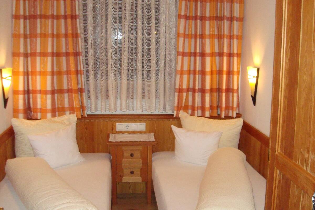 A room with two beds, wooden walls, a window with curtains, and wall-mounted lights.
