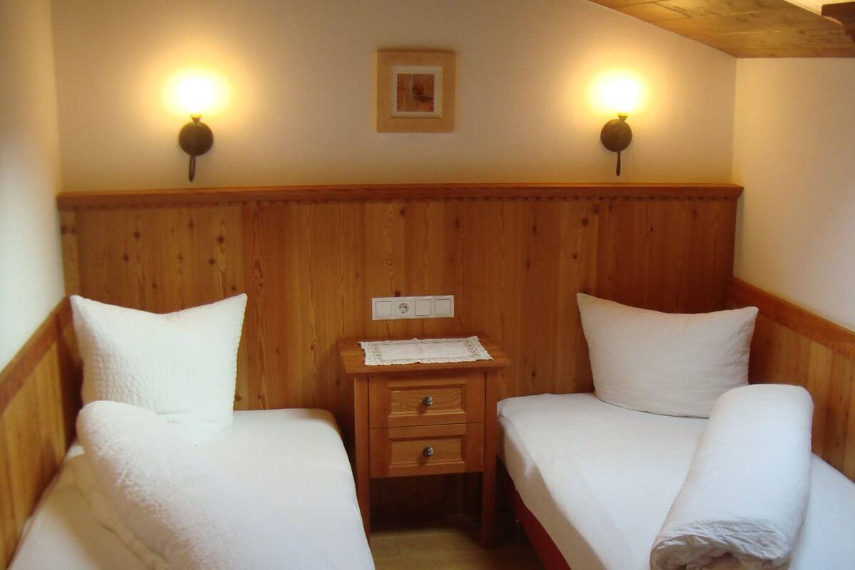 A small, cozy room with two single beds, wooden headboard, a nightstand with a lamp, and a framed picture on the wall.