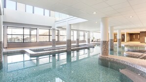 A spacious indoor swimming pool with clear glass walls and a tiled floor, surrounded by white pillars and a high ceiling.