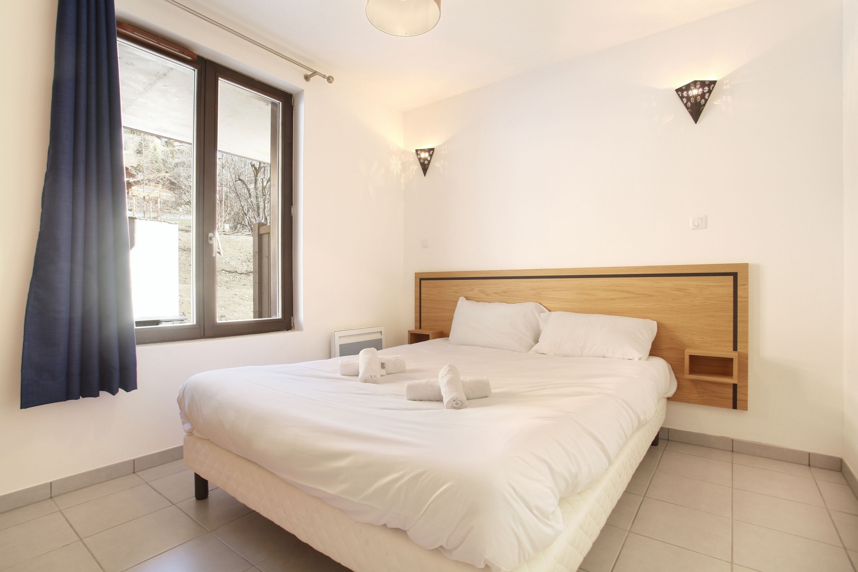 Una cama bien tendida con ropa de cama y toallas blancas, cabecera de madera y una ventana con cortinas.