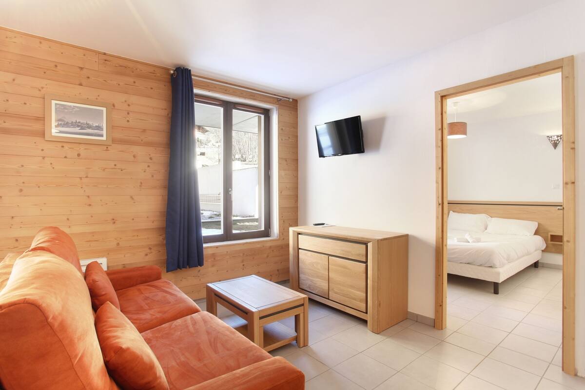 Een moderne kamer met houten vloer, een bank, een tafeltje, een kast, een bed, een televisie en een raam met gordijnen.