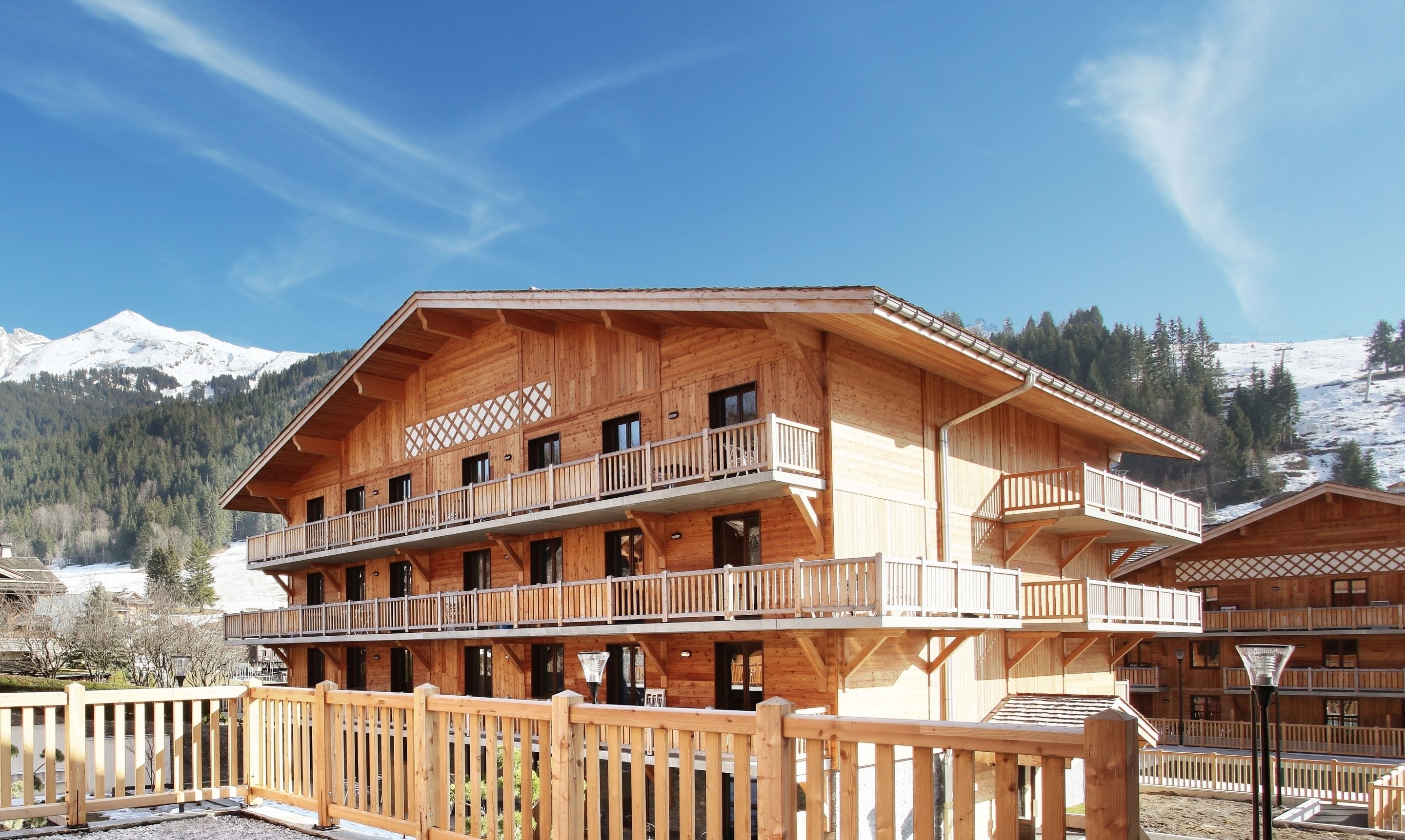 A wooden chalet with multiple balconies, a wooden fence, and a mountainous backdrop.