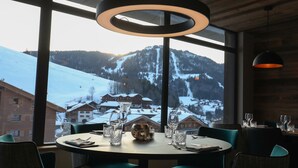 A dining area with a table set for two, large windows offering a view of a snowy mountain landscape, and modern pendant lighting.