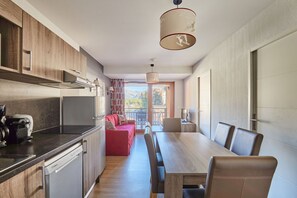 A modern kitchen with a built-in oven, a coffee machine, and a dishwasher. There is a dining area with a wooden table and chairs, and a red sofa. A large window provides a view of the mountains. - Résidence Odalys Aquisana (La Salle-les-Alpes)