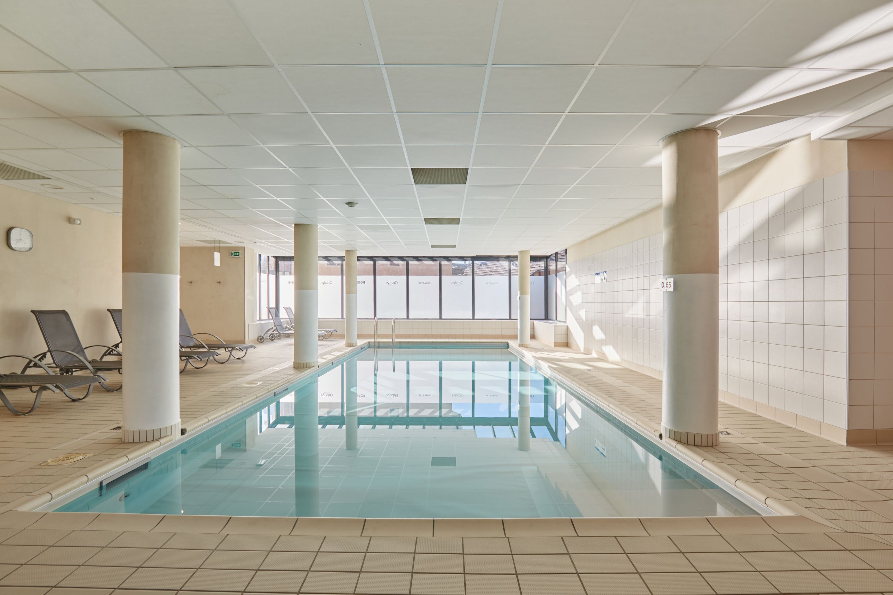 Een binnenzwembad met helder water, omringd door betegelde wanden en zuilen, met ligbedden en een klok aan de muur.
