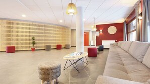 A modern lobby with a large beige sofa, a round table with magazines, red ottomans, and a wooden slatted wall.