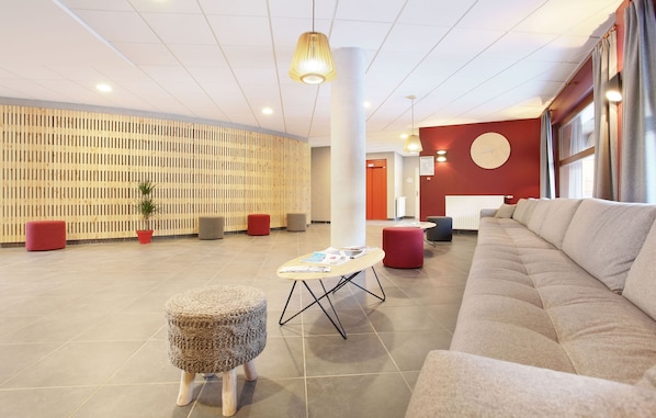A modern lobby with a large beige sofa, a round table with magazines, red ottomans, and a wooden slatted wall. - Résidence Odalys Aquisana (La Salle-les-Alpes)